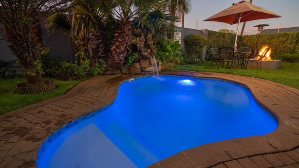 A swimming pool with a waterfall feature, surrounded by a brick patio and lush greenery.