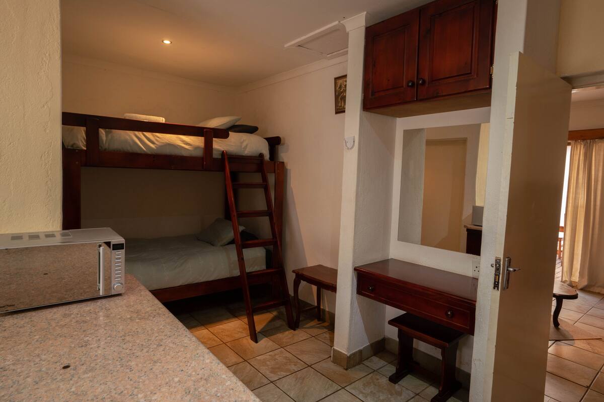A compact room with a bunk bed, a desk, a chair, a microwave, and a door leading to another room.