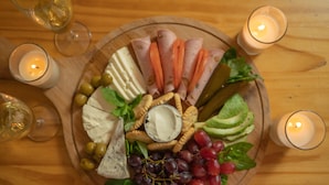 A wooden board with a variety of cheeses, meats, fruits, and crackers, surrounded by lit candles on a wooden table.