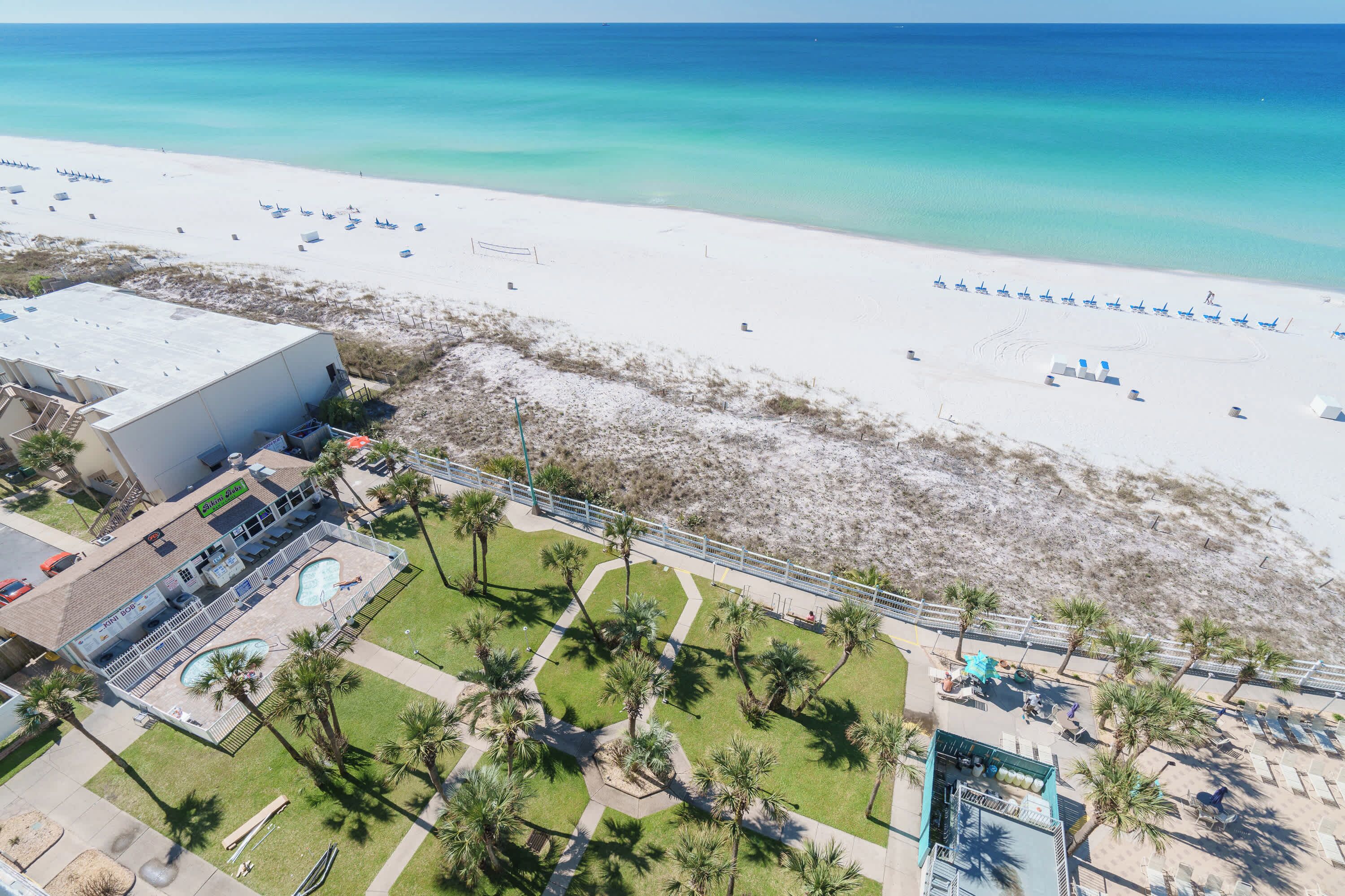 Vista aérea de un complejo turístico con piscina, palmeras y sillas de playa.