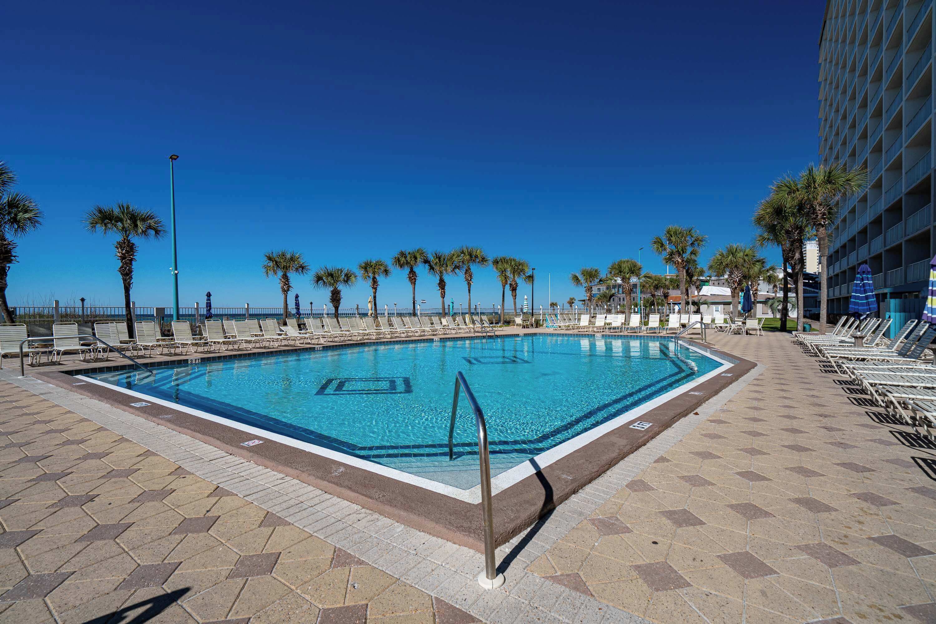 Una piscina con solárium de baldosas, palmeras y sillas de descanso.