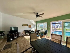 A living room with a TV, wicker furniture, a ceiling fan, and a view of a swimming pool.