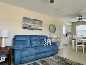 A living room with a blue sofa, a dining table with chairs, a kitchen area, and a ceiling fan.
