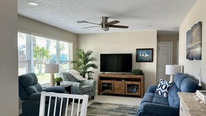 Ein Wohnzimmer mit Fernseher, Sitzgelegenheiten, Deckenventilator und Blick durch große Fenster.