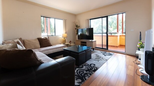 A modern living room with a sofa, a coffee table, a flat-screen TV, and a sliding glass door leading to a balcony. - Sydney Two-Level 3BDR 3BATH near CBD & Olympic Park  (Burwood)