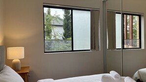 A hotel room with a bed, a nightstand with a lamp, a large window with a view of greenery, and a mirror reflecting the room. - Sydney Two-Level 3BDR 3BATH near CBD & Olympic Park  (Burwood)