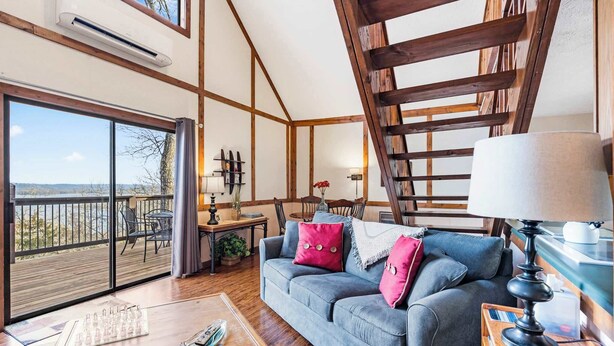 A cozy living room with a large window, a wooden staircase, and a balcony with a view. - Skipjack Cottage Branson (Branson West)