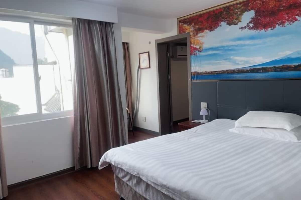 Ein Hotelzimmer mit einem Bett, einem Fenster mit Vorhängen, einem Gemälde einer Berglandschaft und einer Tür zu einem anderen Raum.