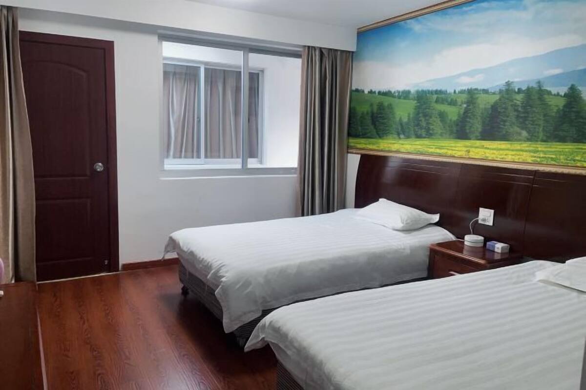 Ein Hotelzimmer mit zwei Betten, einem Fenster mit Vorhängen, einem Landschaftsbild und einer Holztür.