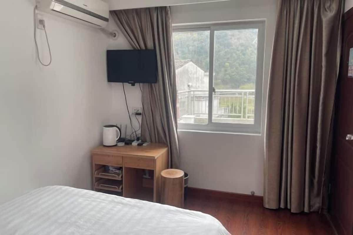 Ein Schlafzimmer mit einem Bett, einem Holztisch, einem Fernseher, einem Fenster mit Vorhängen und Blick auf ein Gebäude.
