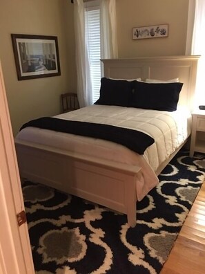 Une chambre à coucher avec un lit, une table de chevet, une fenêtre avec des stores, un tableau encadré au mur et un tapis à motifs.