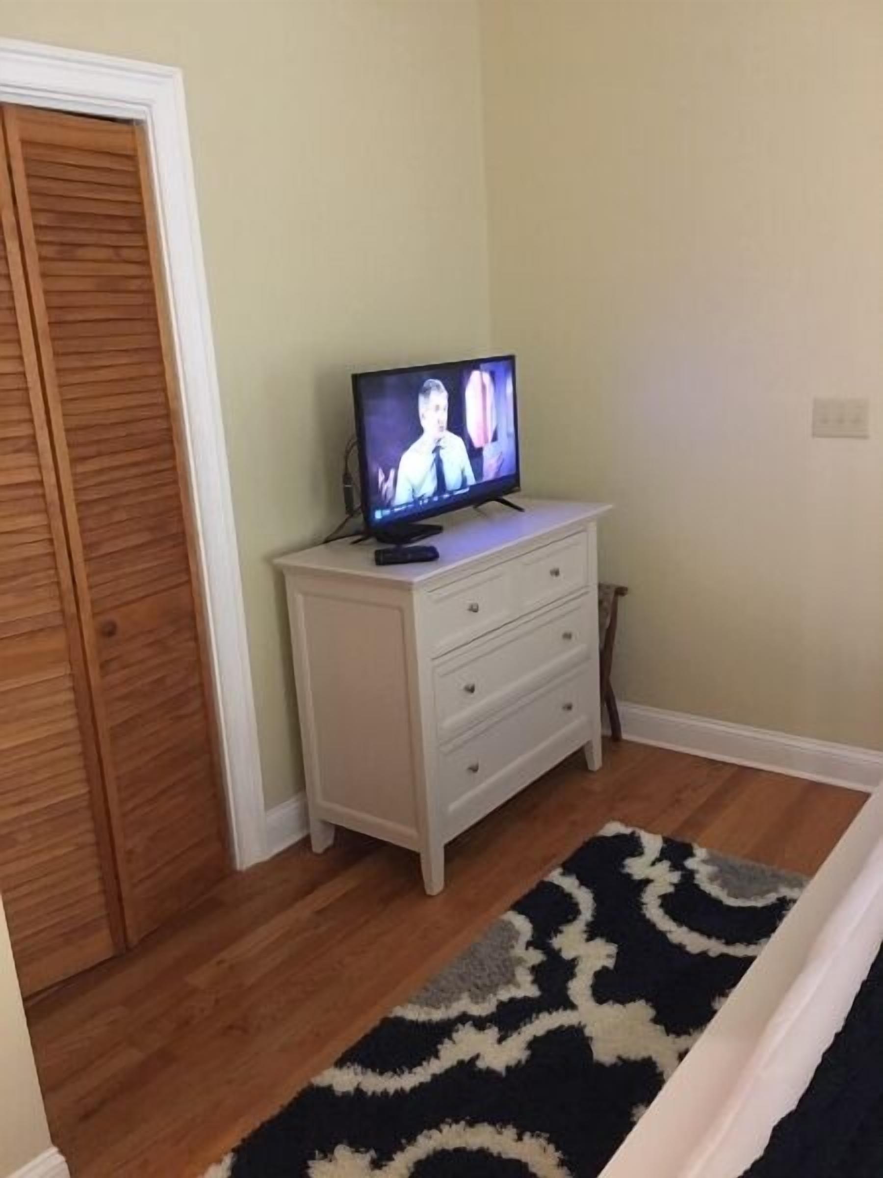 A room with a television on a white dresser, a wooden door, and a patterned rug on the floor.