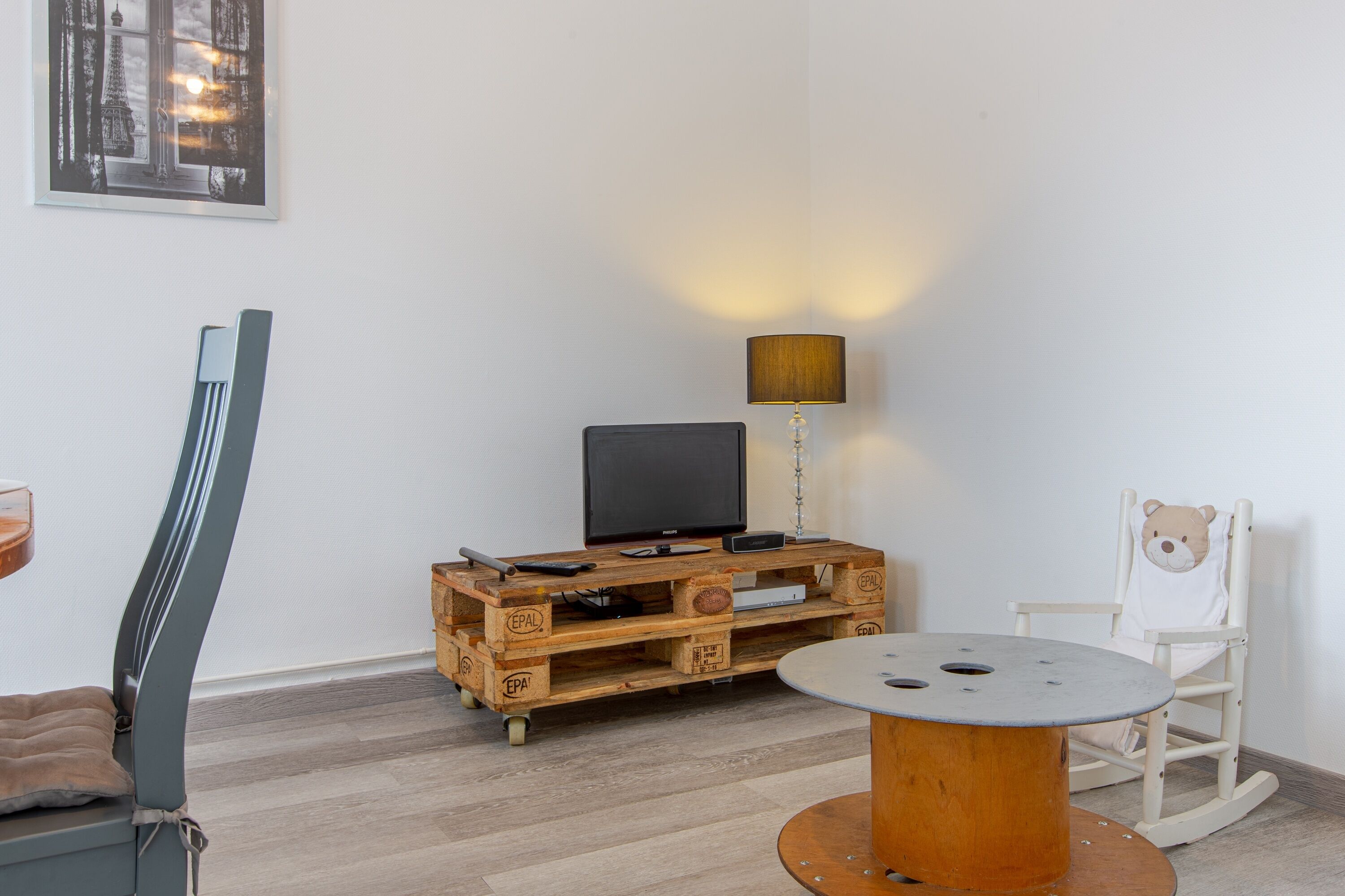 A room with a wooden TV stand, a lamp, a round table, a rocking chair, and a framed picture on the wall.