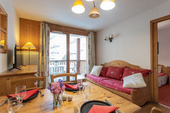 Une table à manger en bois avec des chaises, un canapé avec des coussins rouges et une fenêtre avec des rideaux.