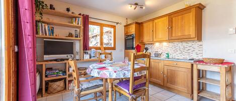 A cozy kitchen with wooden cabinets, a dining table with chairs, a TV, and a window with curtains.