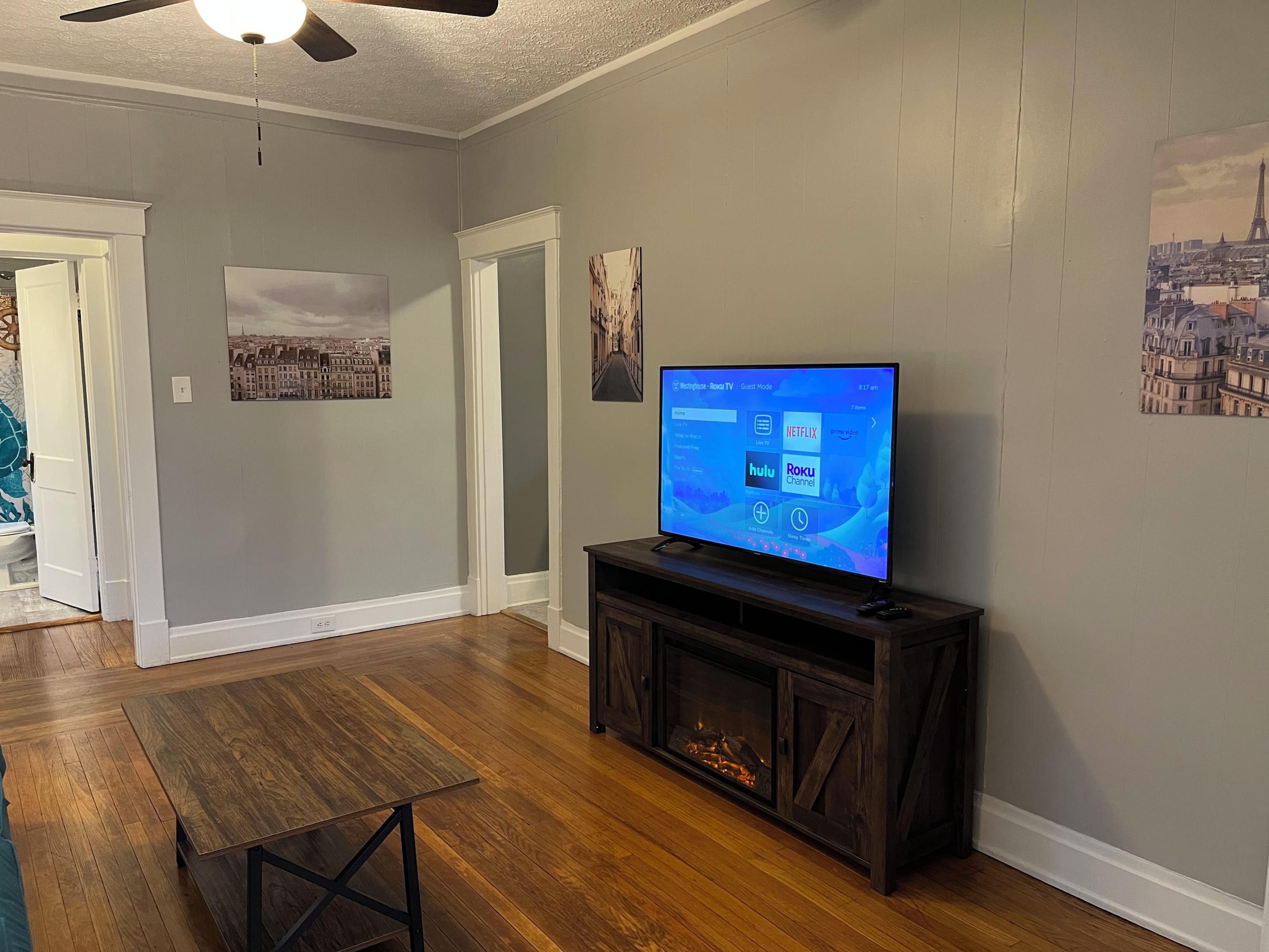 Una sala de estar con un televisor de pantalla plana sobre un soporte de madera, una chimenea y una puerta que da a otra habitación.