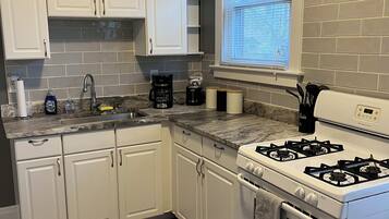 Una cocina con gabinetes blancos, estufa a gas, cafetera y ventana con cortinas estampadas.