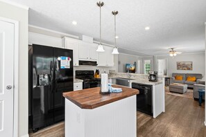 A modern kitchen with a central island, stainless steel appliances, pendant lighting, and a dining area with a gray sofa and orange cushions. - 🏝️Close to Beach-Pet Friendly-Private Home-3BR Beachin' Fun (Navarre)