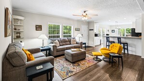 Ein Wohnzimmer mit einer Couch, einem Hocker und einem Sessel, im Hintergrund eine Küche und ein Deckenventilator.