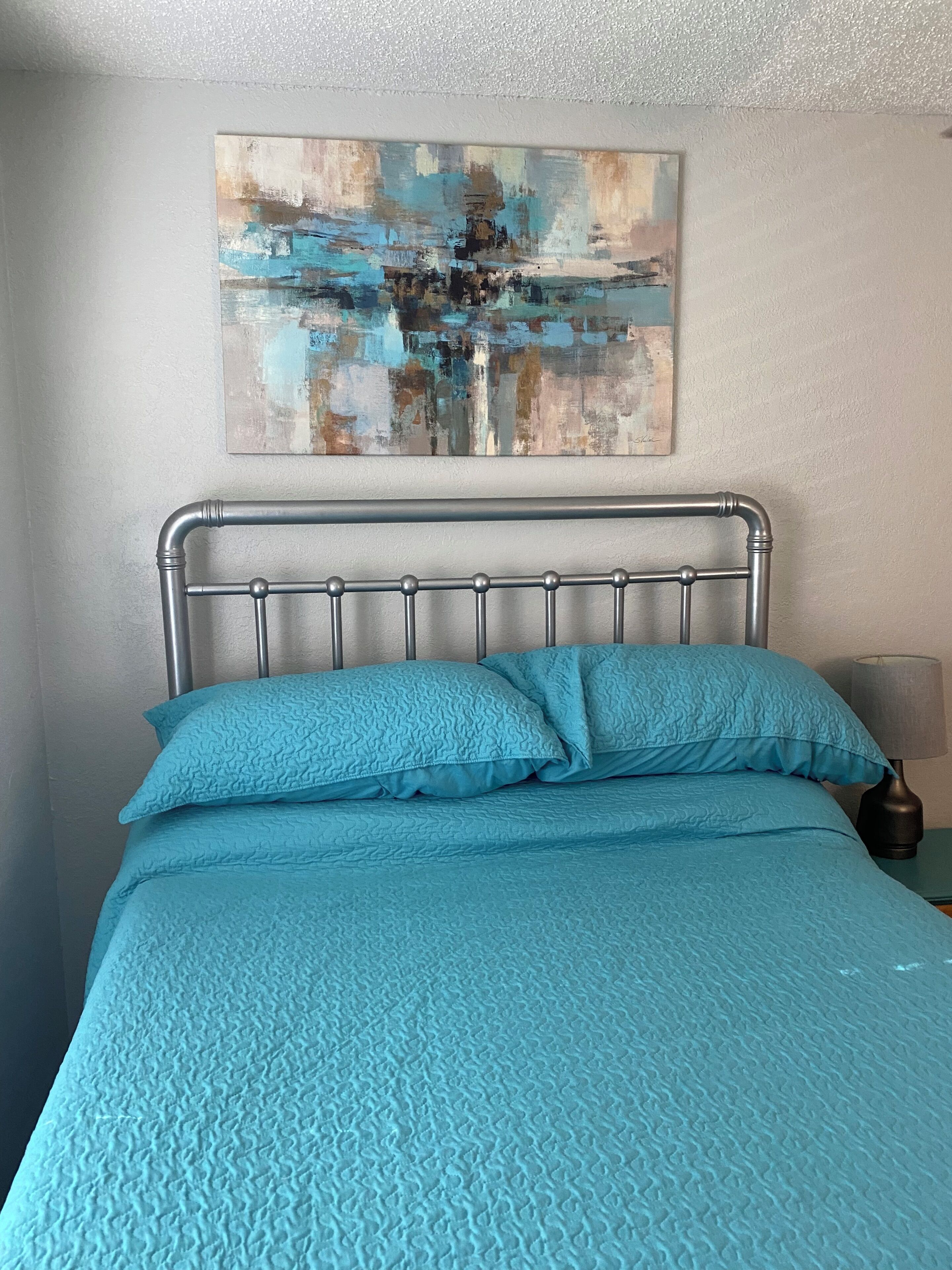 A bed with a metal headboard, a blue quilted bedspread, and a painting on the wall.