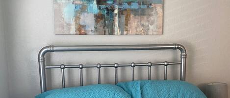A bed with a metal headboard, a blue quilted bedspread, and a painting on the wall.