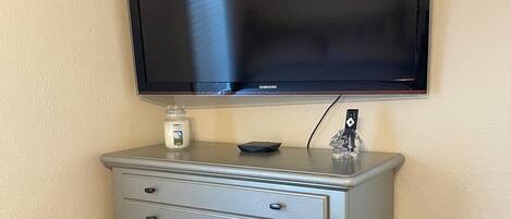 Ein Fernseher ist über einer grauen Kommode mit Schubladen angebracht, obenauf befindet sich ein kleines Objekt und seitlich ein Mülleimer.