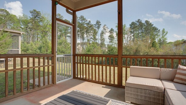 A covered outdoor deck with a wicker sofa and a bench, overlooking a wooded area.
