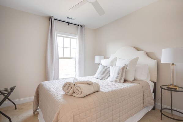A neatly made bed with rolled towels, a bedside lamp, and a window with curtains. - Stylish & Centrally Located Home (Cary)