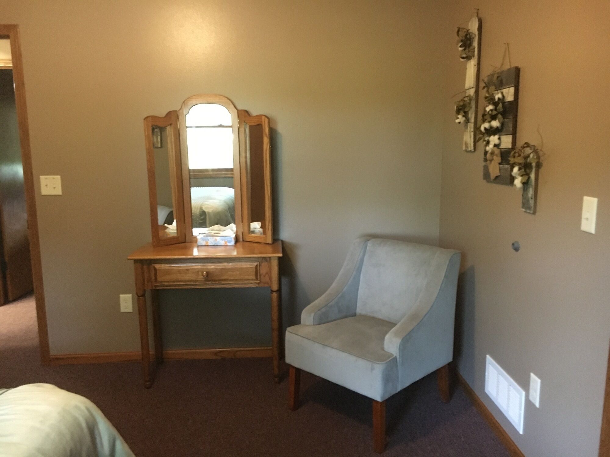 Een slaapkamer met een houten dressoir, een lichtblauwe fauteuil, een spiegel en een wanddecoratie.