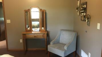 Een slaapkamer met een houten dressoir, een lichtblauwe fauteuil, een spiegel en een wanddecoratie.