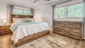 A bedroom with a bed, wooden dresser, nightstands, a ceiling fan, and large windows. - Taste of Tuscany - Hot Tub, Fire Pit & Pool Table (Fredericksburg)