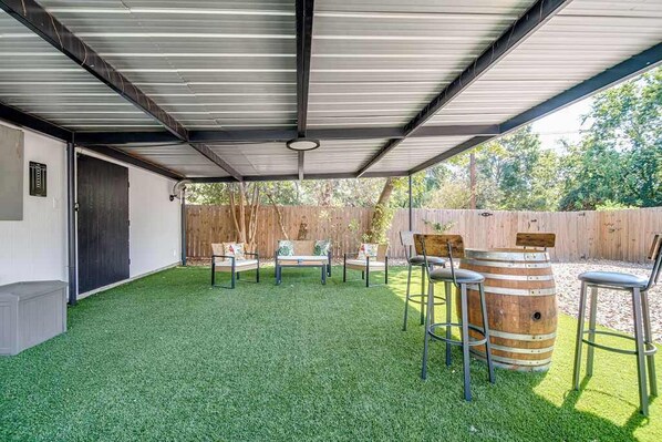 Een buitenpatio met kunstgras, metalen stoelen, een houten vat en een overdekt gedeelte met een metalen dak.