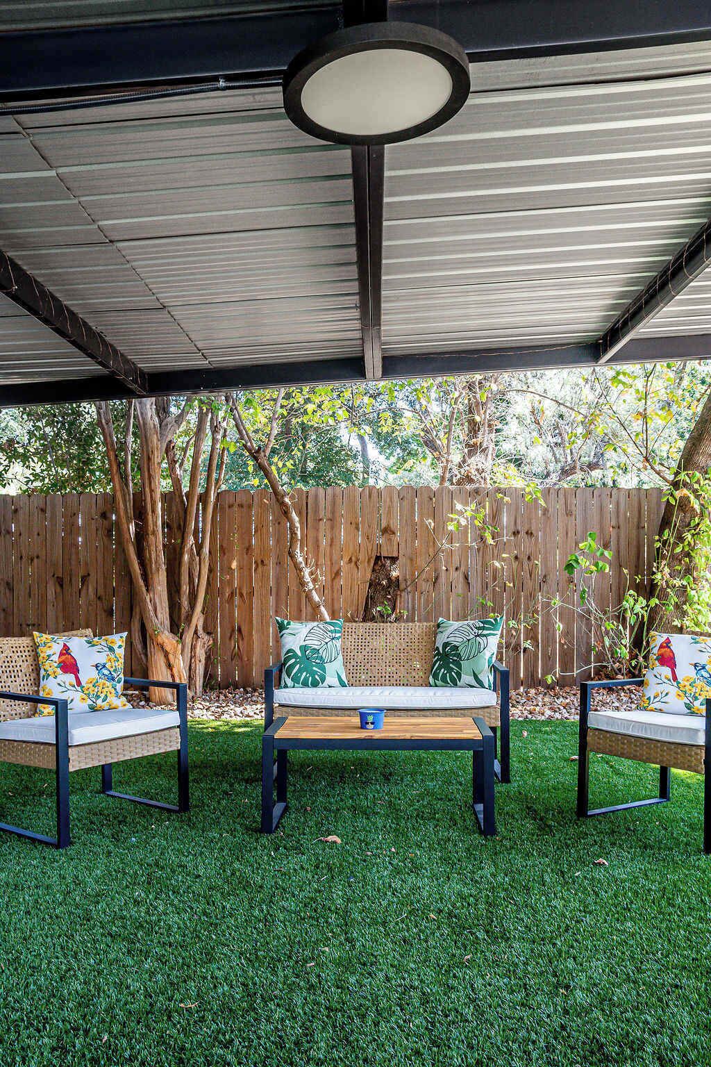 A covered patio with a wooden table and two chairs, surrounded by artificial grass.