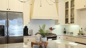 A modern kitchen with a stainless steel refrigerator, white cabinets, a marble countertop, and a central island with a potted plant and candle. - The Sadie, 3BR Townhome by VelvetDitchVillas (Oxford)