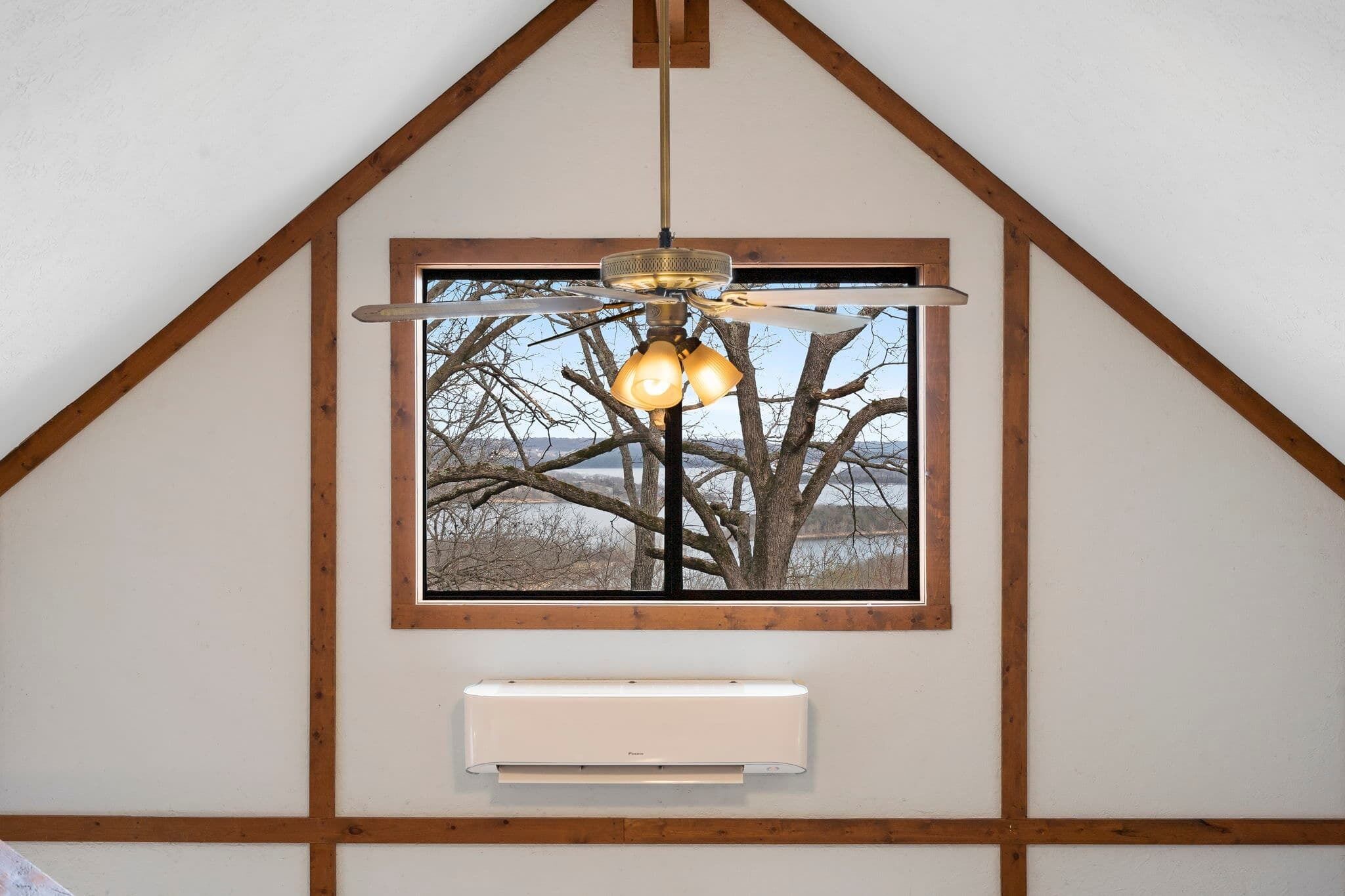 A room with a sloped ceiling, a ceiling fan, a window with a view of bare trees, and a wall-mounted air conditioner.
