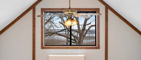 A room with a sloped ceiling, a ceiling fan, a window with a view of bare trees, and a wall-mounted air conditioner.