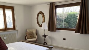 A bedroom with a large bed, a chair, a bedside table, a lamp, a mirror, and a window with curtains.