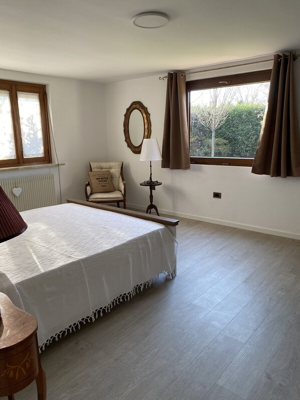 A bedroom with a large bed, a chair, a bedside table, a lamp, a mirror, and a window with curtains.