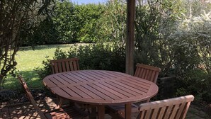 A wooden outdoor patio with a round table and chairs, surrounded by lush greenery and a clear sky.