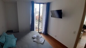 Habitación de hotel con cama, televisor montado en la pared y vista de la ciudad a través de un ventanal grande.