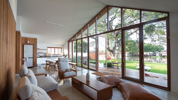 Ein modernes Wohnzimmer mit großen Fenstern, einer Küche im Hintergrund und Blick auf einen Garten und Wasser.