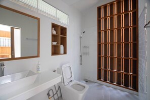 A bathroom with a white sink, a toilet, a shower, and a wooden shelf unit. - River house for relaxation and remote work station (nakornpathom)