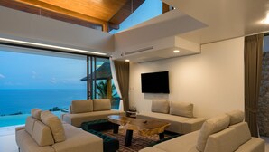 Un salon moderne doté d’une grande fenêtre donnant sur une plage, meublé de canapés beige et d’une table basse en bois.