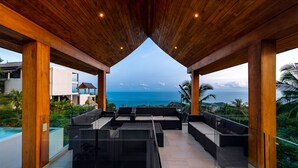 Un espace de détente extérieur doté de poutres en bois et de rampes en verre, offrant une vue sur une piscine et l’océan.
