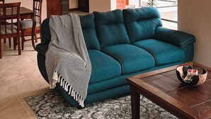 A living room with a teal sofa, a wooden coffee table, a patterned rug, a dining area with chairs, and a kitchen visible through an archway. - Casa Lia  (San Isidro)
