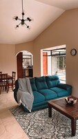 Uma sala de estar com sofá azul-turquesa, mesa de centro de madeira, tapete estampado, área de jantar com cadeiras e cozinha visível através de um arco.