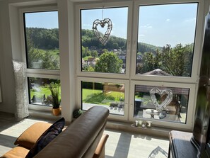 A room with a large window overlooking a green landscape, a cushioned bench, and a decorative heart hanging from the window.