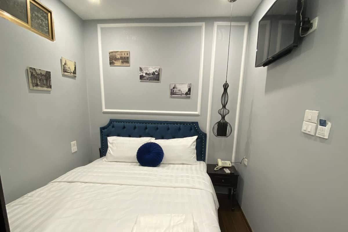 A neatly made bed with white linens and a blue accent pillow, a bedside table with a lamp, and a wall-mounted TV.