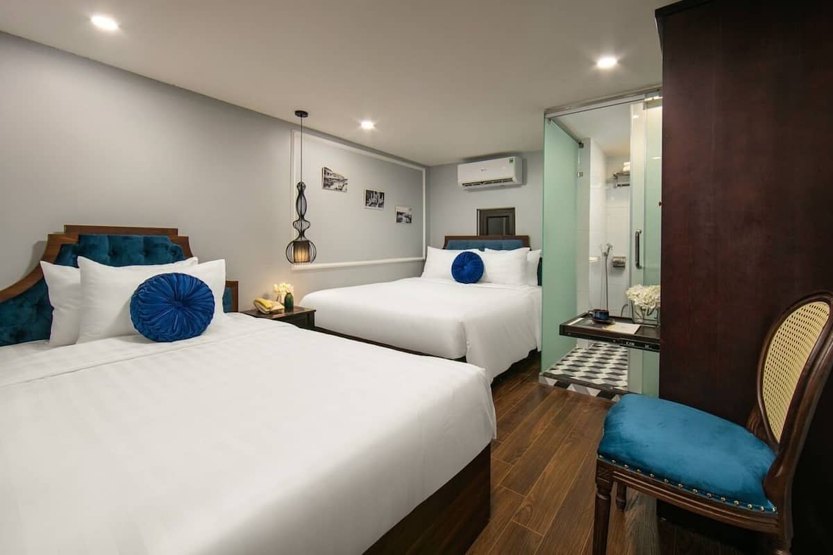 A hotel room with two beds, a small table, a chair, and a bathroom visible through an open door.