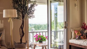 Une pièce avec un banc en bois, une plante en pot, un canapé agrémenté de coussins à motifs floraux et une vue sur la verdure, visible à travers une porte vitrée.